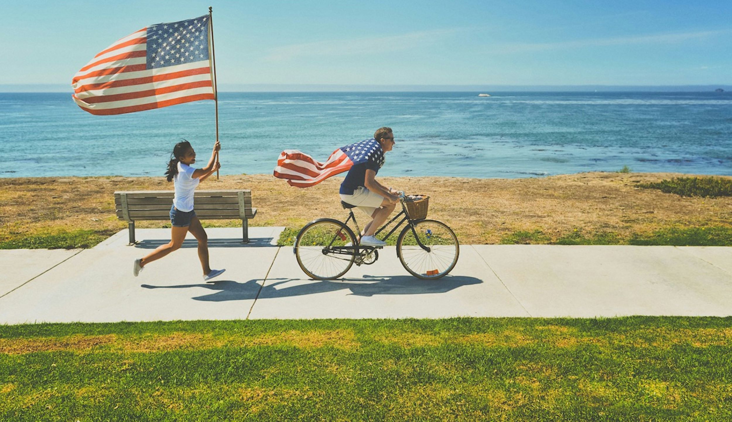 A woman runs with a American flag, while a man rides a bike with the American Flag draped around his shoulders.