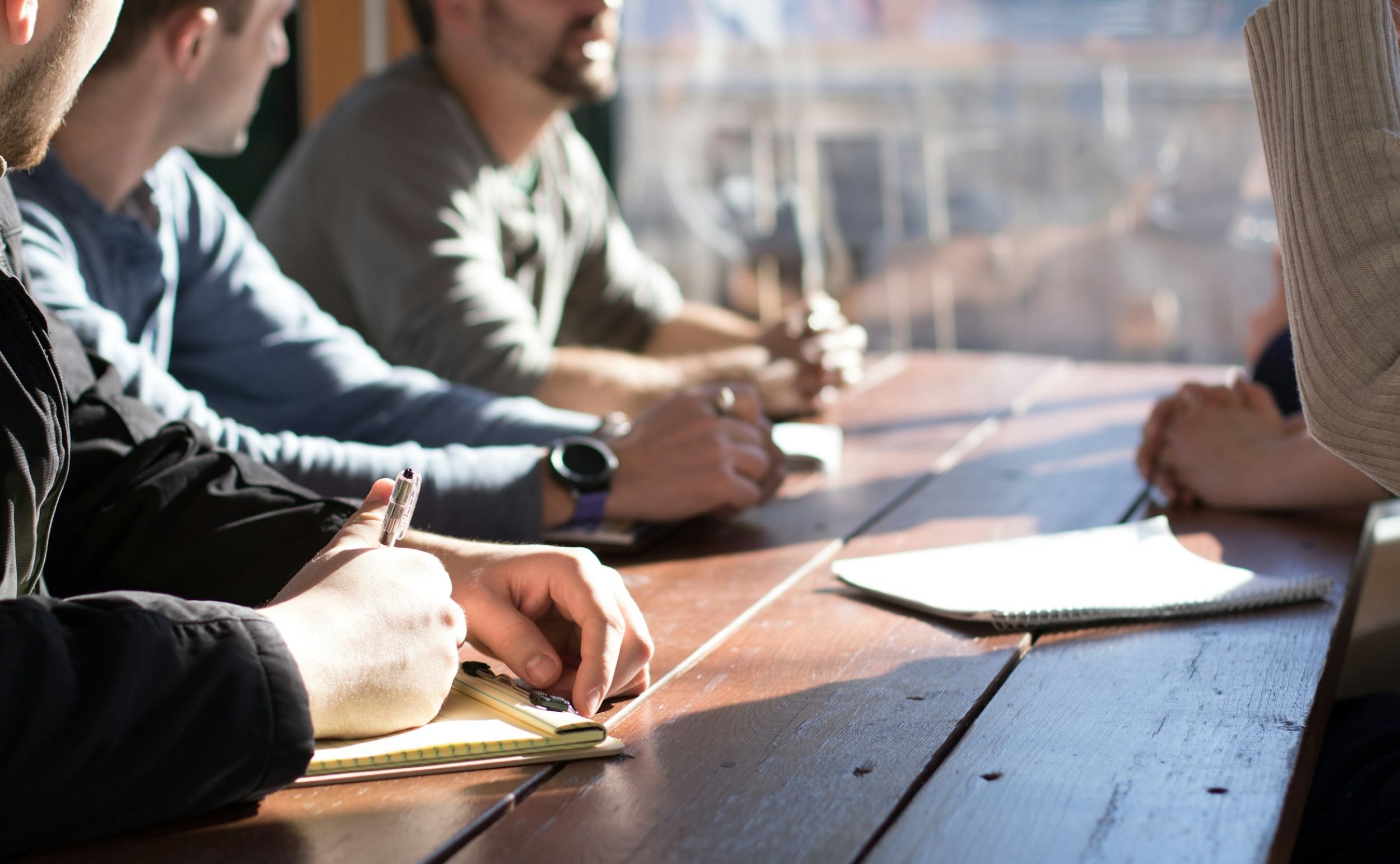 A group of men talk at a wooden table with a notepad and pen in hand.