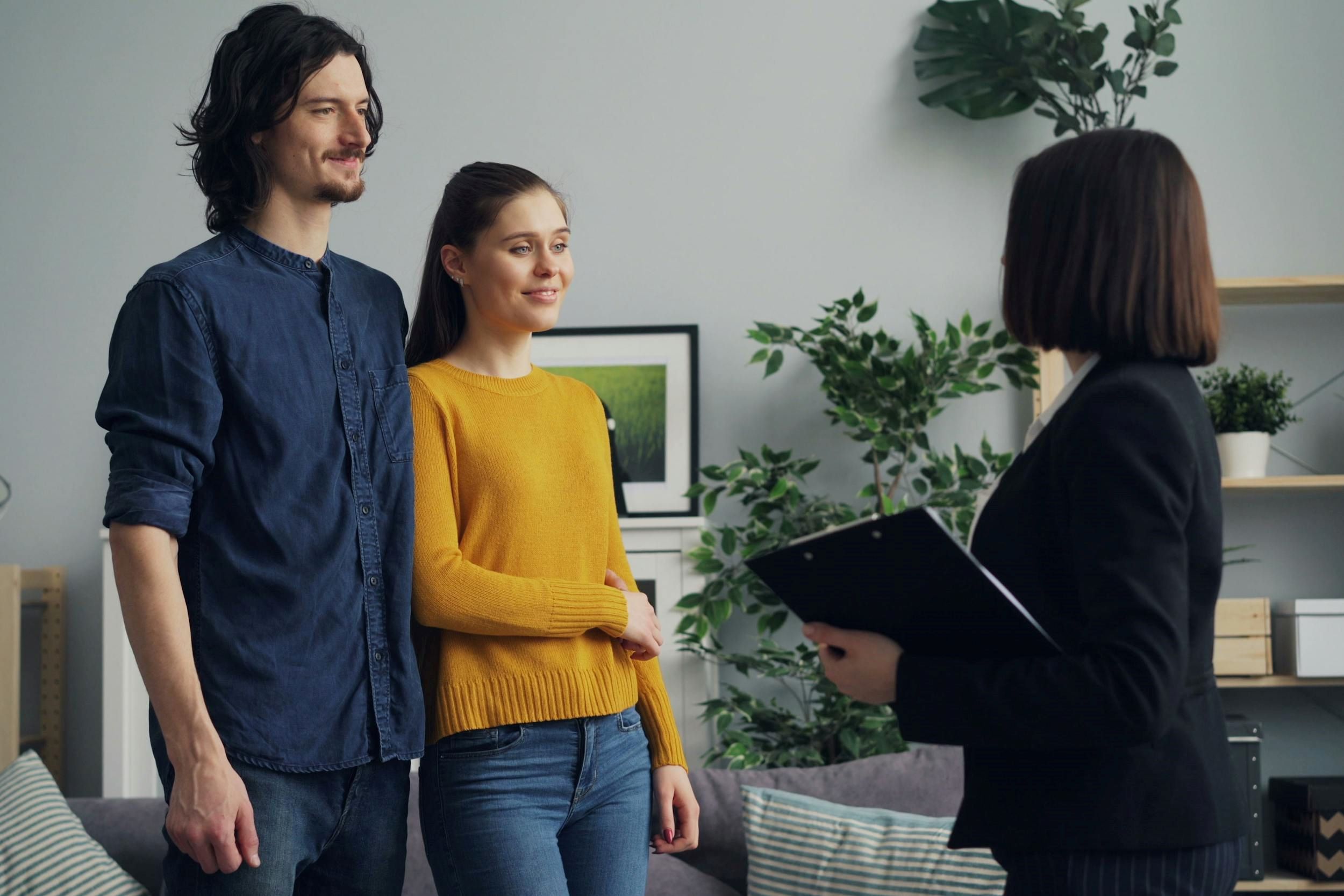 An agent speaks with a couple that stands near a grey couch.