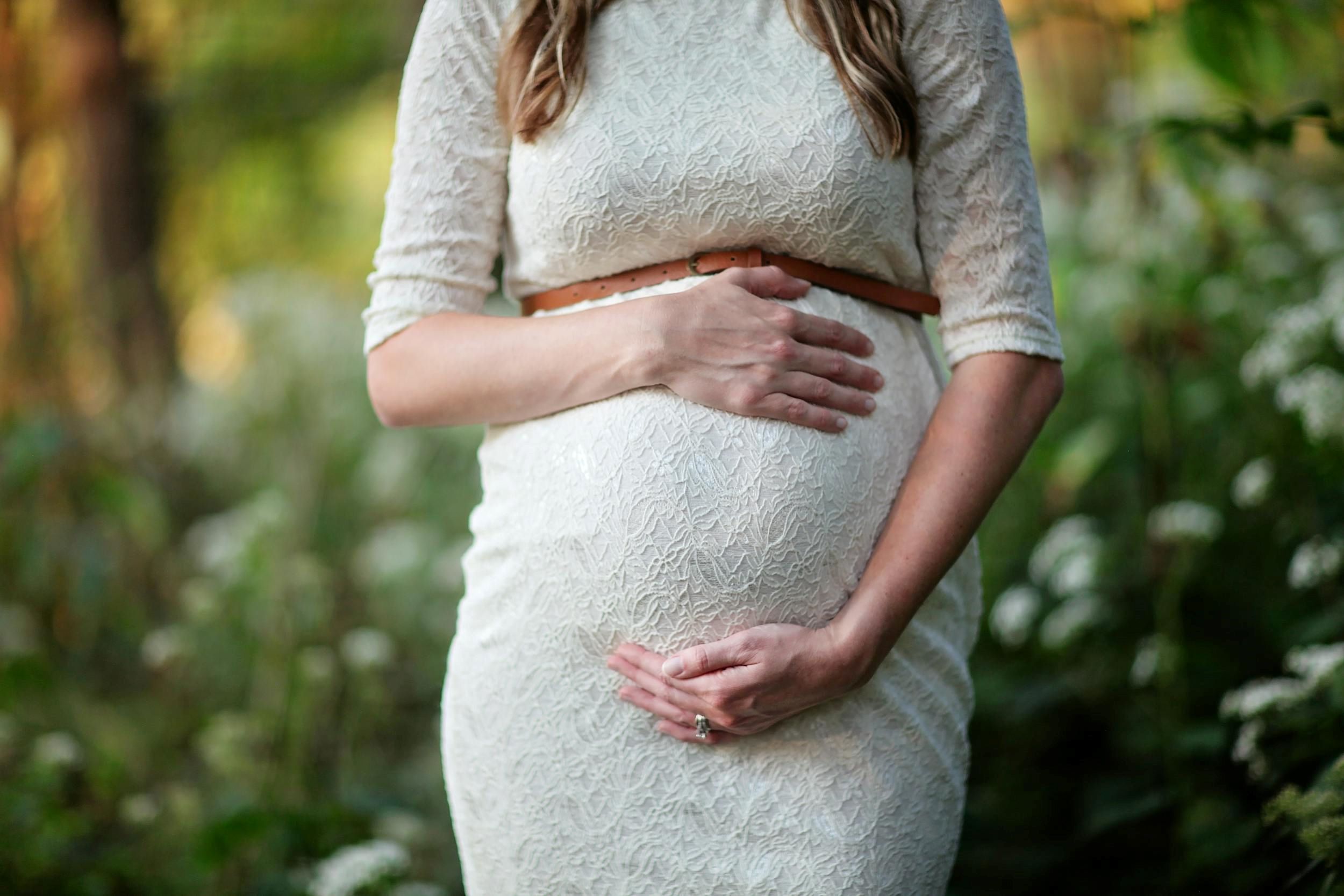 A pregnant woman touches her stomach in front of a cluster of trees and bushes.