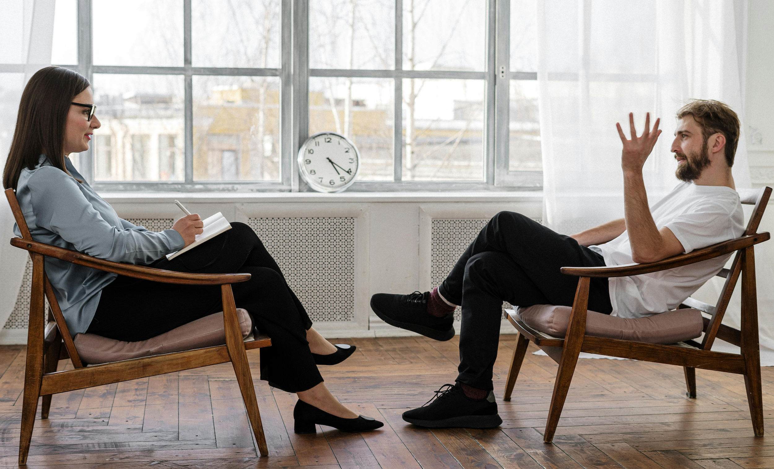 A therapist speaks with her patient in front of a large window with white curtains.