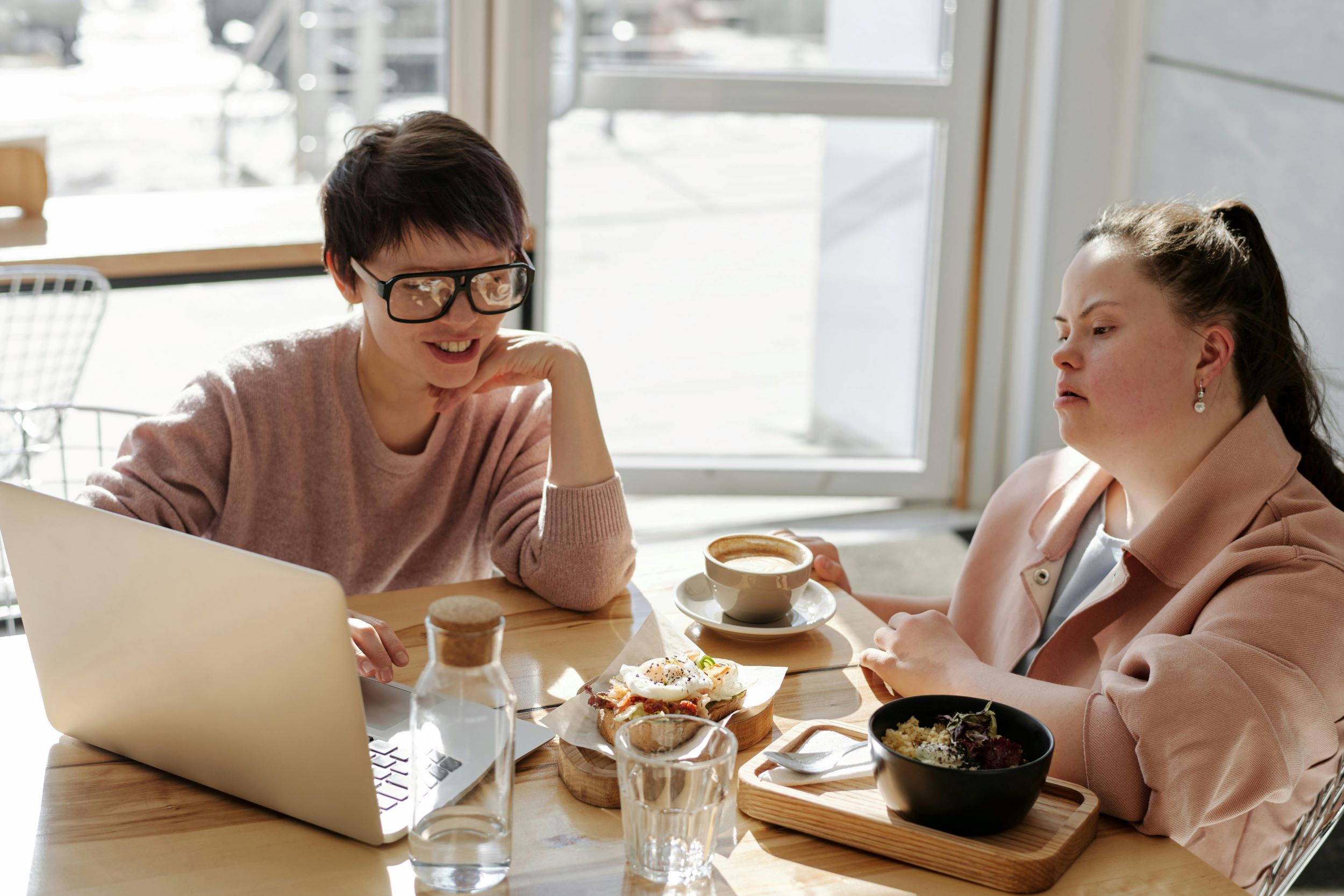 Two young women sit in a well-lit space and stare at a computer.