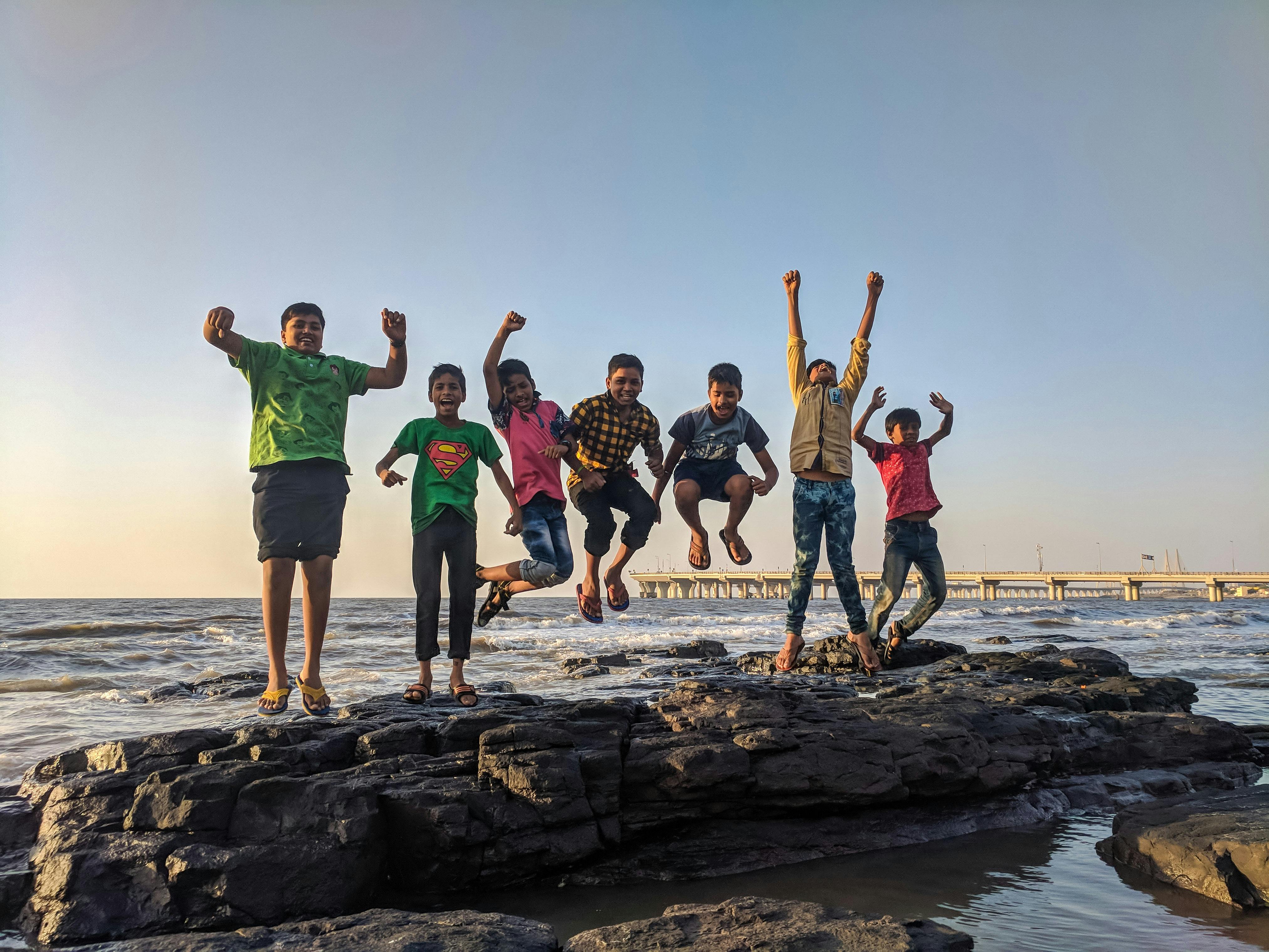 Seven children laugh and jump together on a wide, flat rock on the beach. 