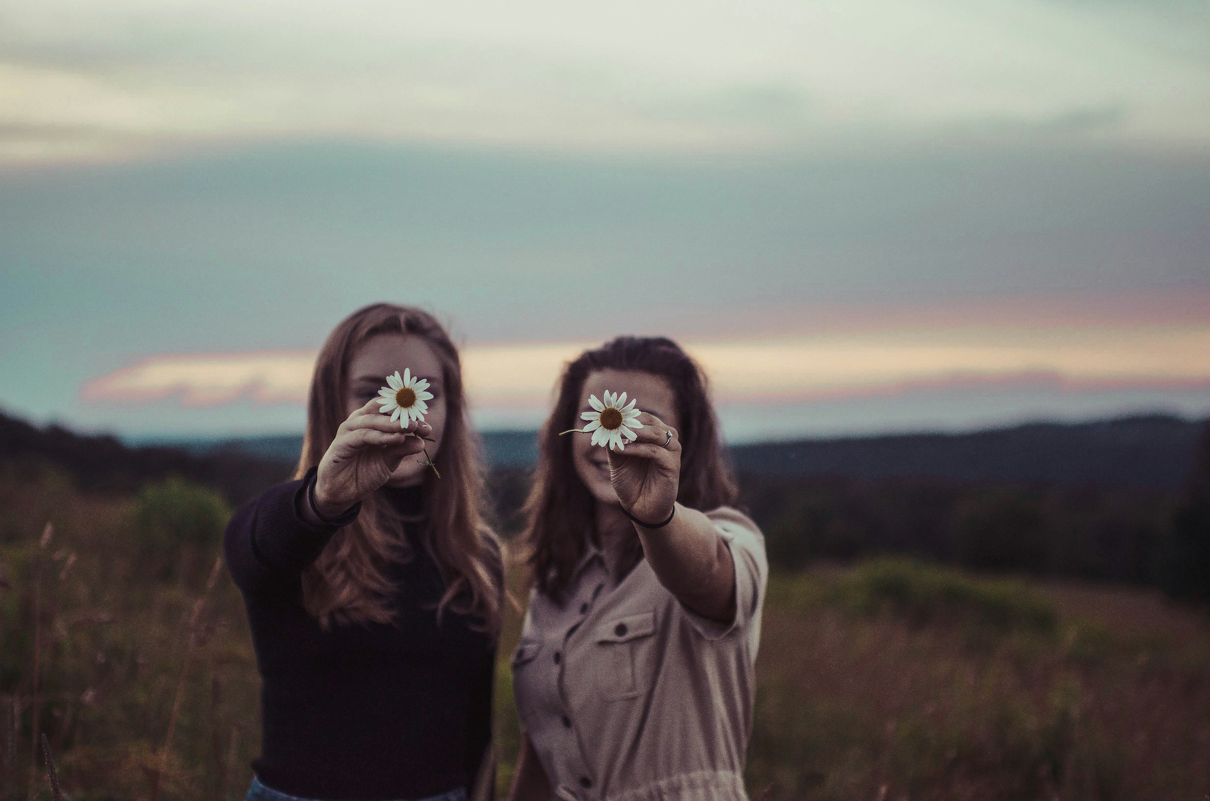 Two women smile while holding flowers in front of a small meadow.