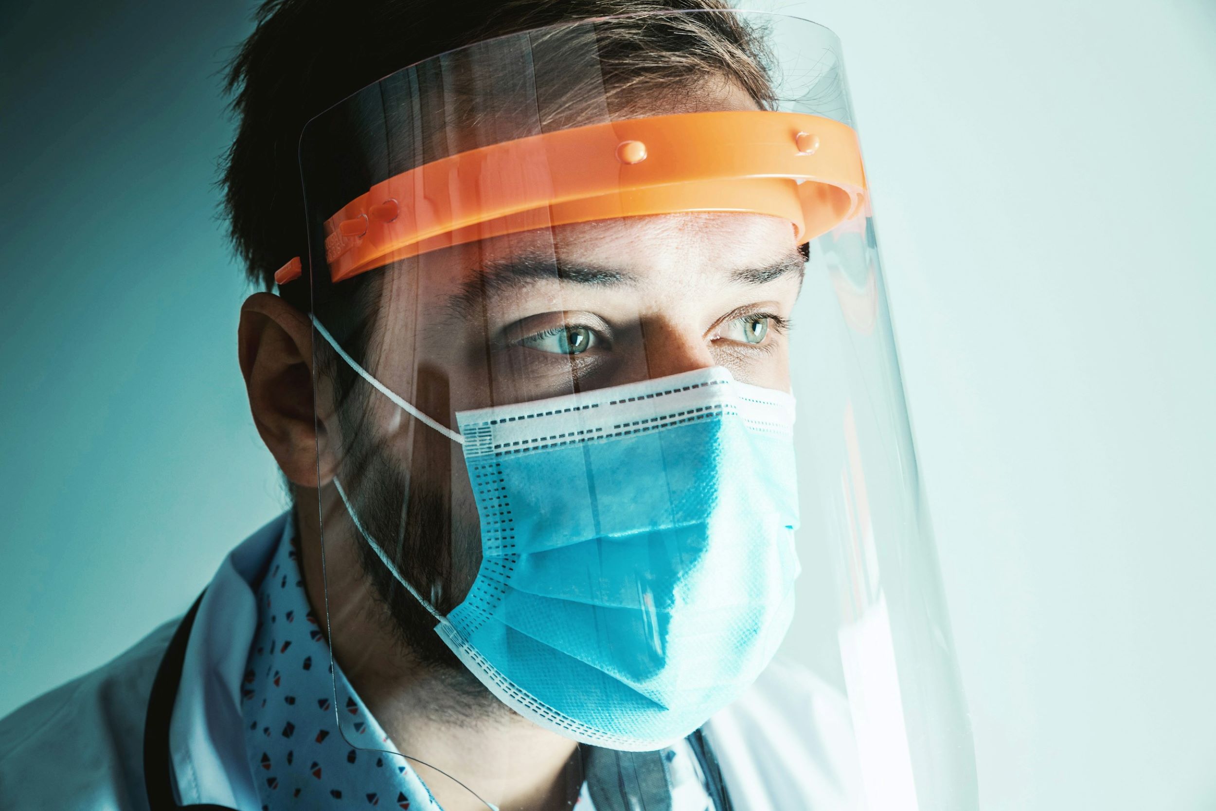A doctor wears a face shield and mask in front of a white and gray backdrop.