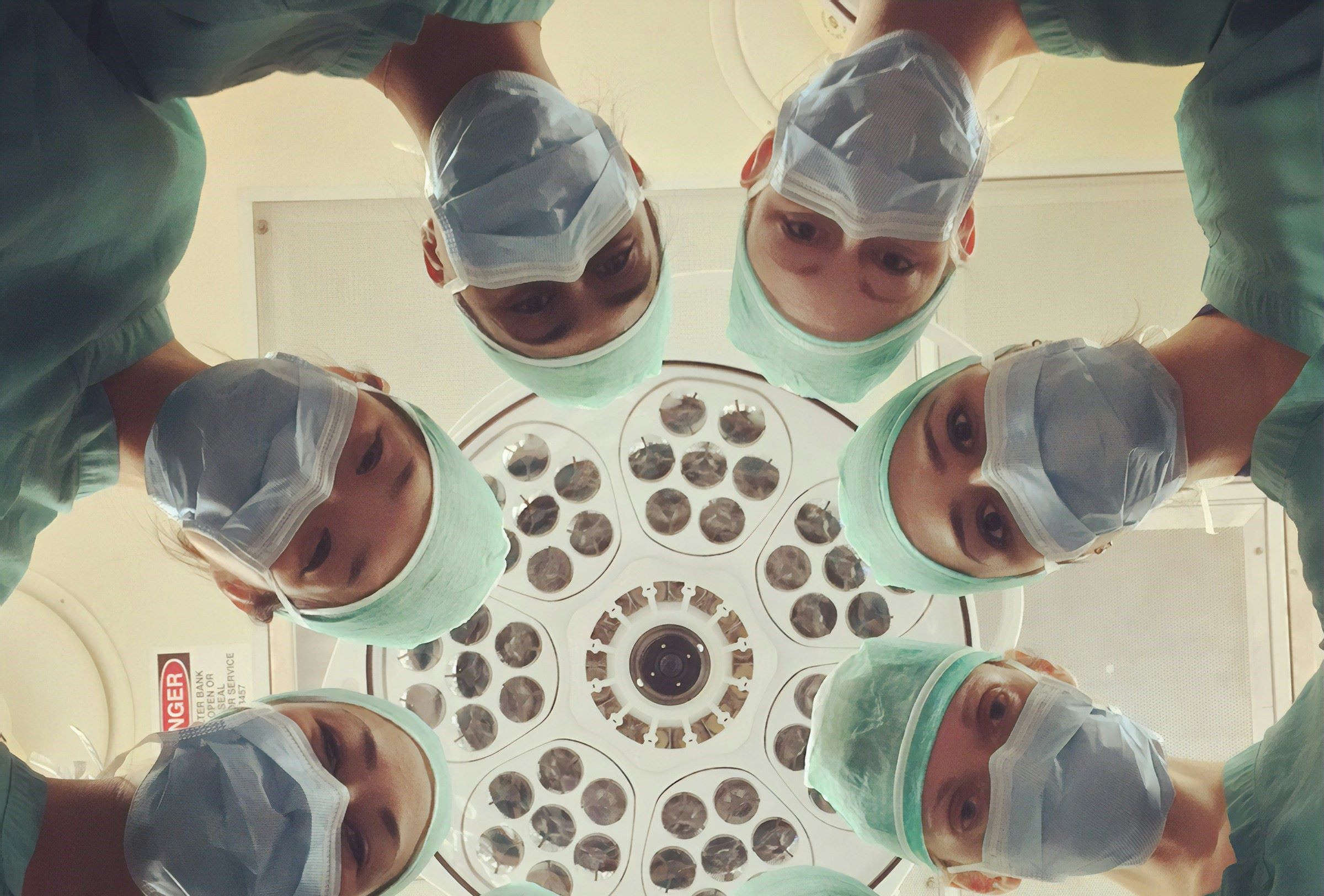 Six doctors stare at a patient with the ceiling and overhead light as their backdrop.