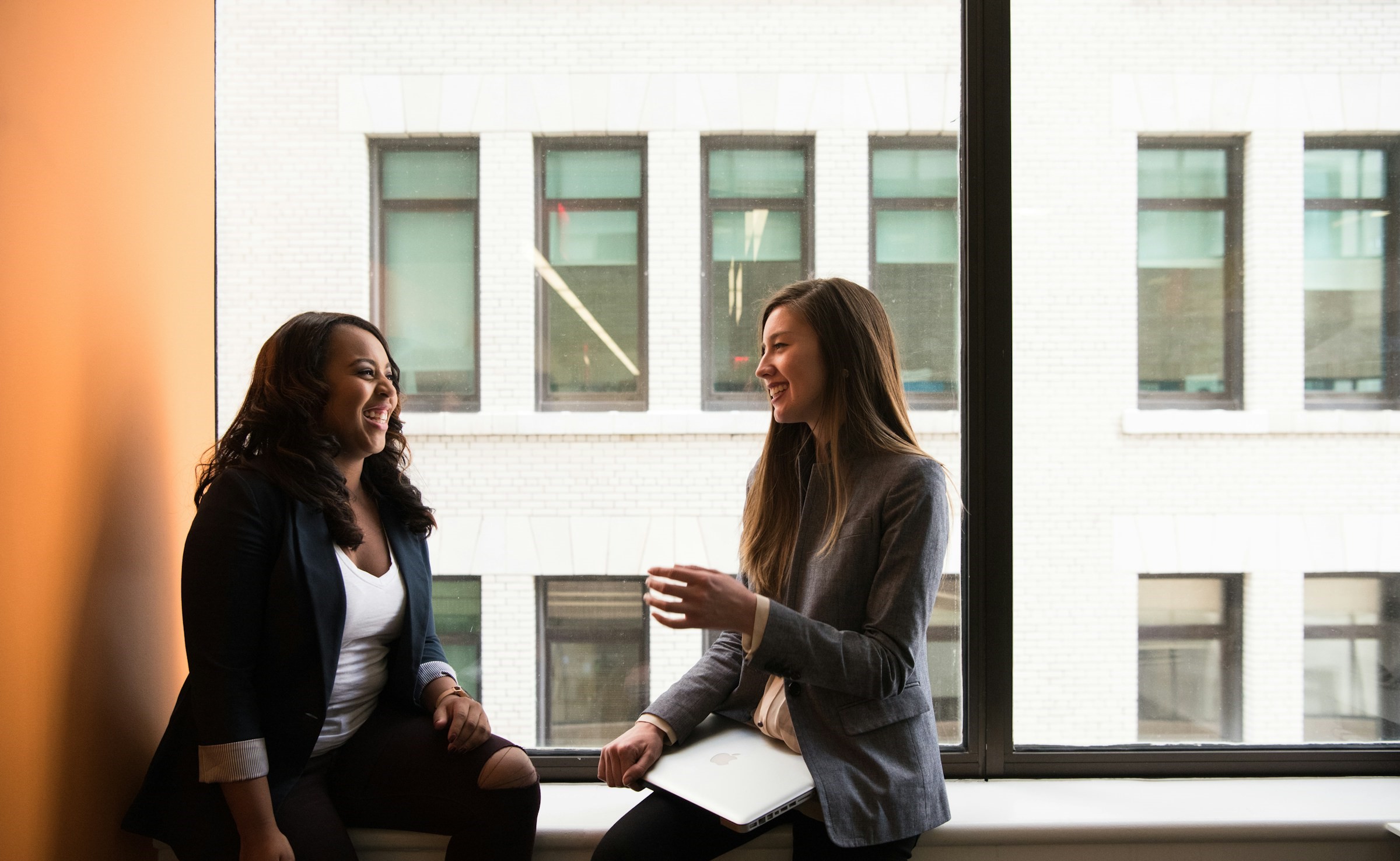 Two women speak near a window.