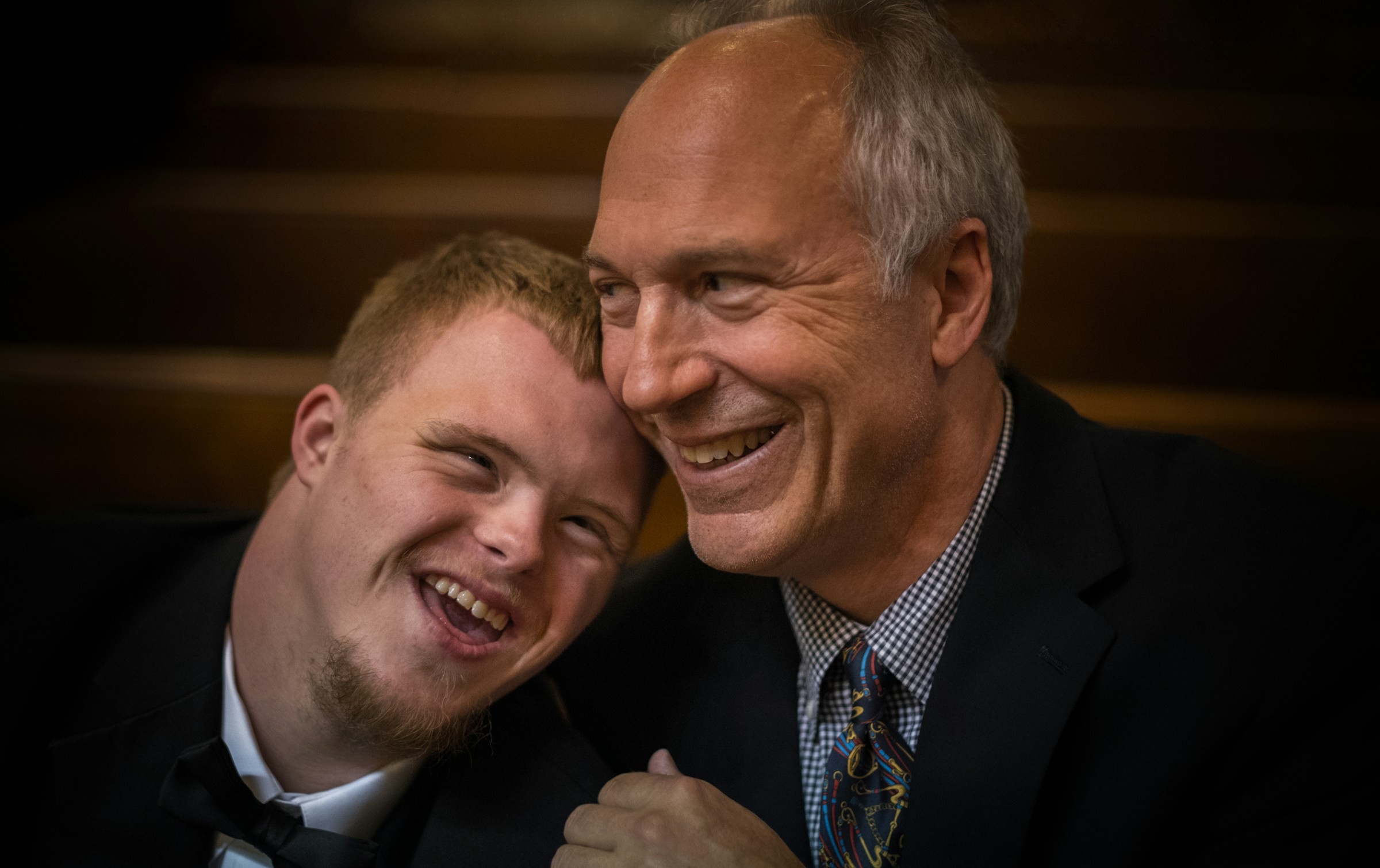 A man with Down Syndrome smiles and leans on his father's shoulder.