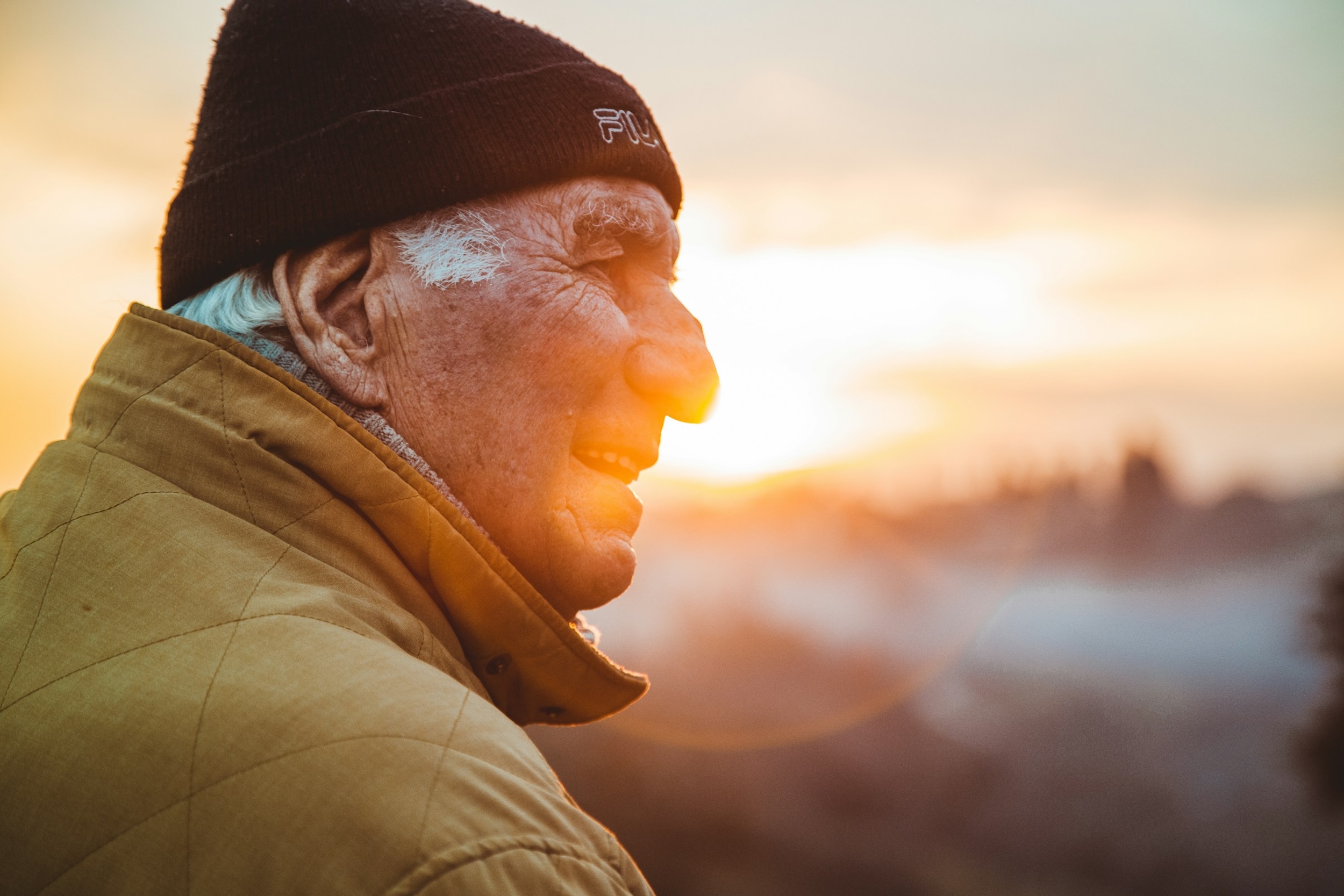 An elderly man smiles warmly as sunlight flares behind him, casting a golden glow.