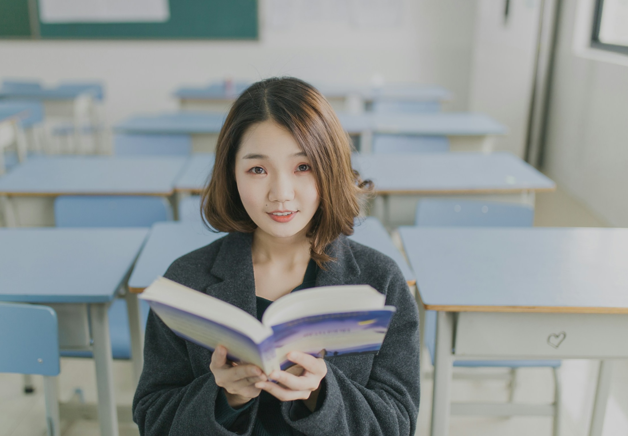 A woman stands in a classroom, smiling warmly as she holds a book in her hands.