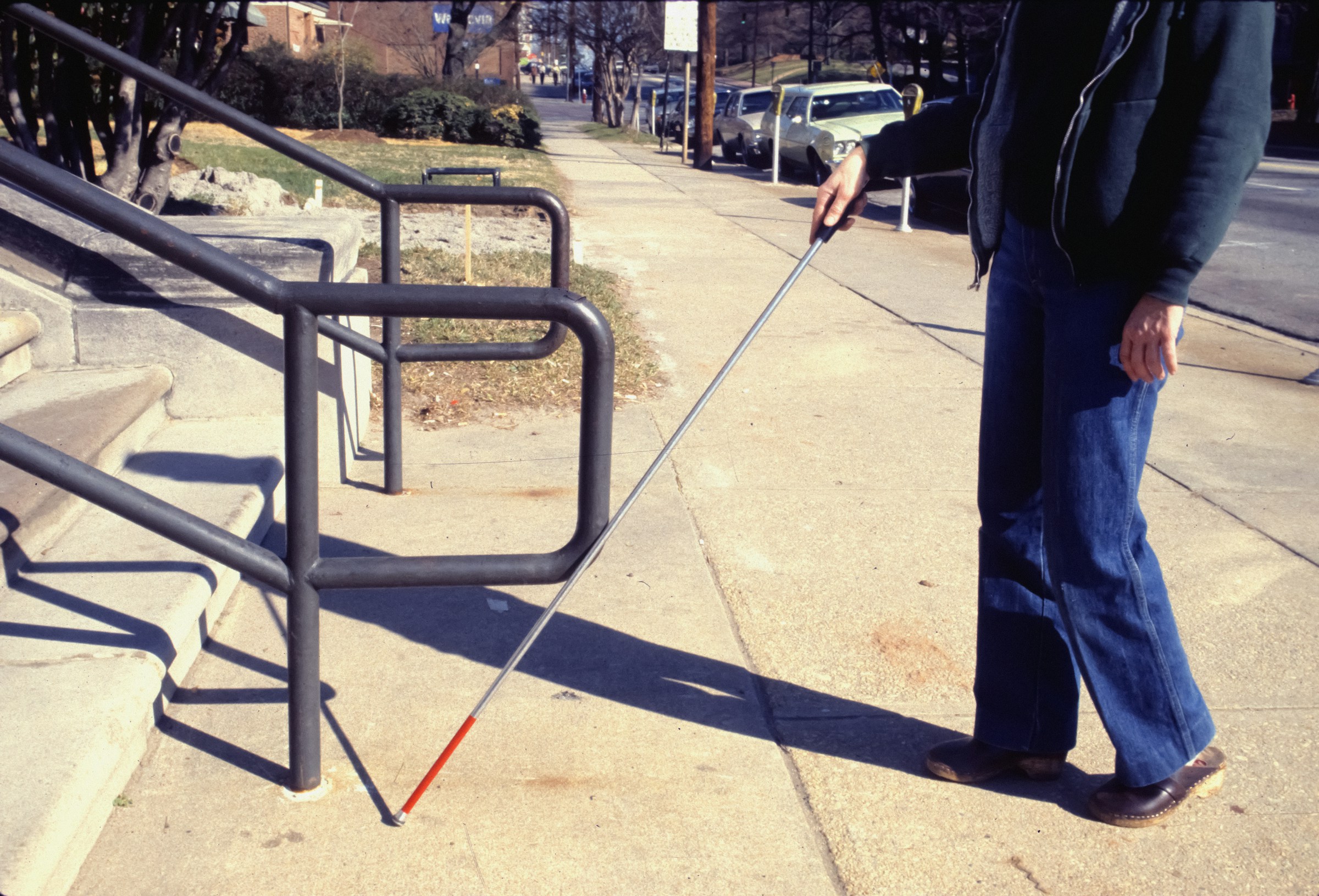 A blind man clutches his walking stick and places it in front of a staircase.
