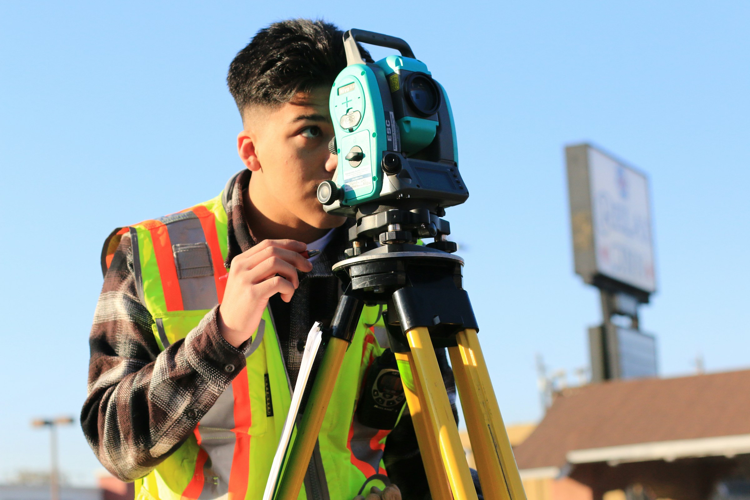 A construction worker wears a bright yellow vest and peers through a device.