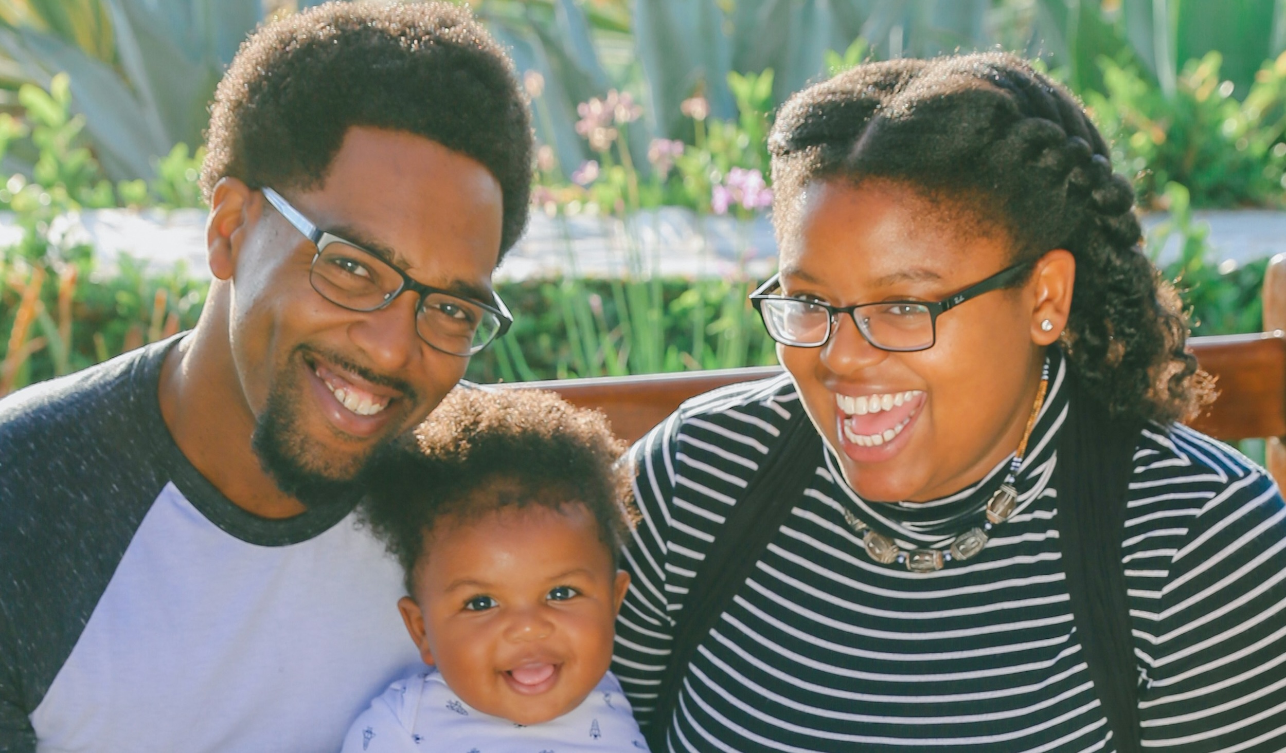 Two parents smile with their baby with flowers as their backdrop.