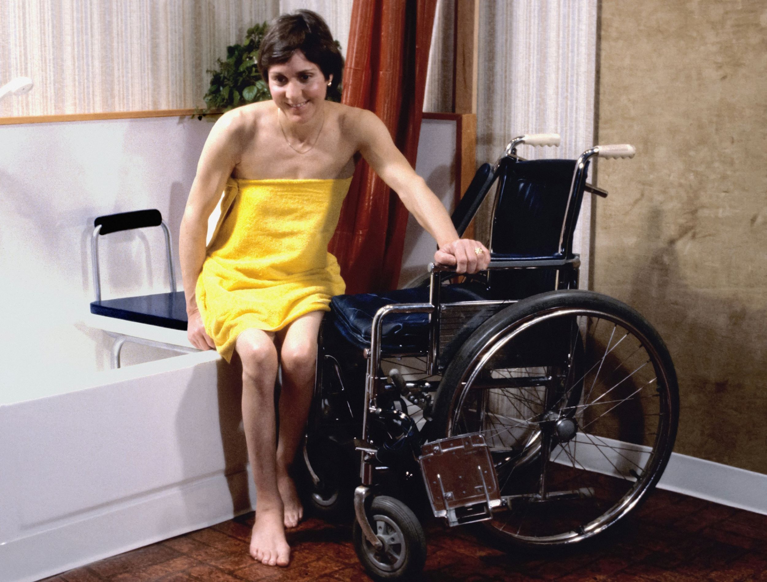 A woman in a yellow dress smiles warmly as she rests her hands on her wheelchair.