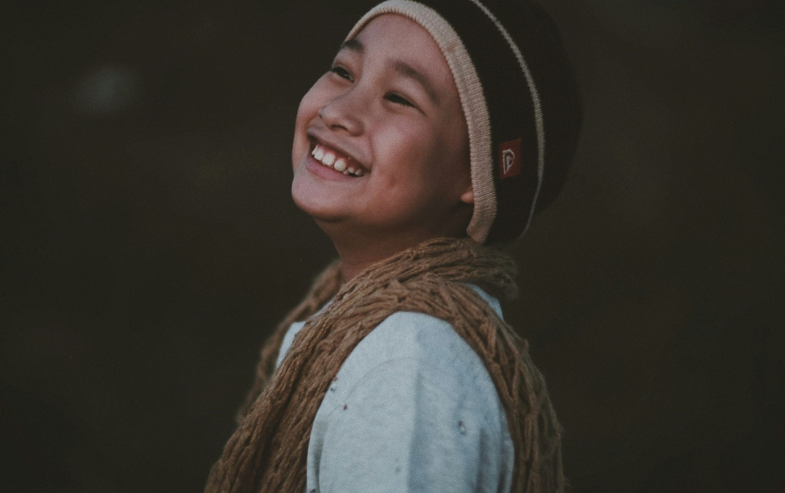 A child smiles in front of a dark brown backdrop.