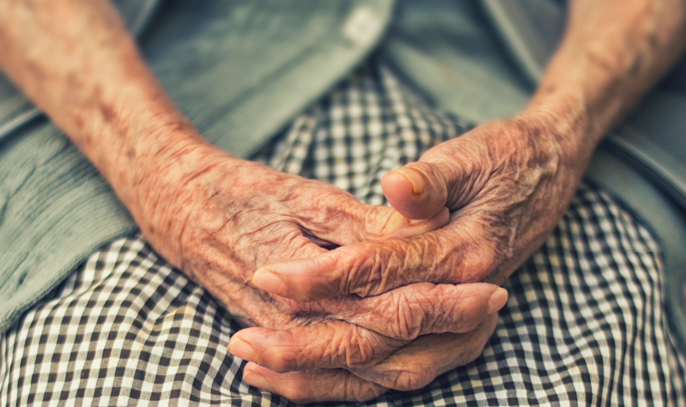 An elderly woman rests her clasped hands gently on her plaid skirt.