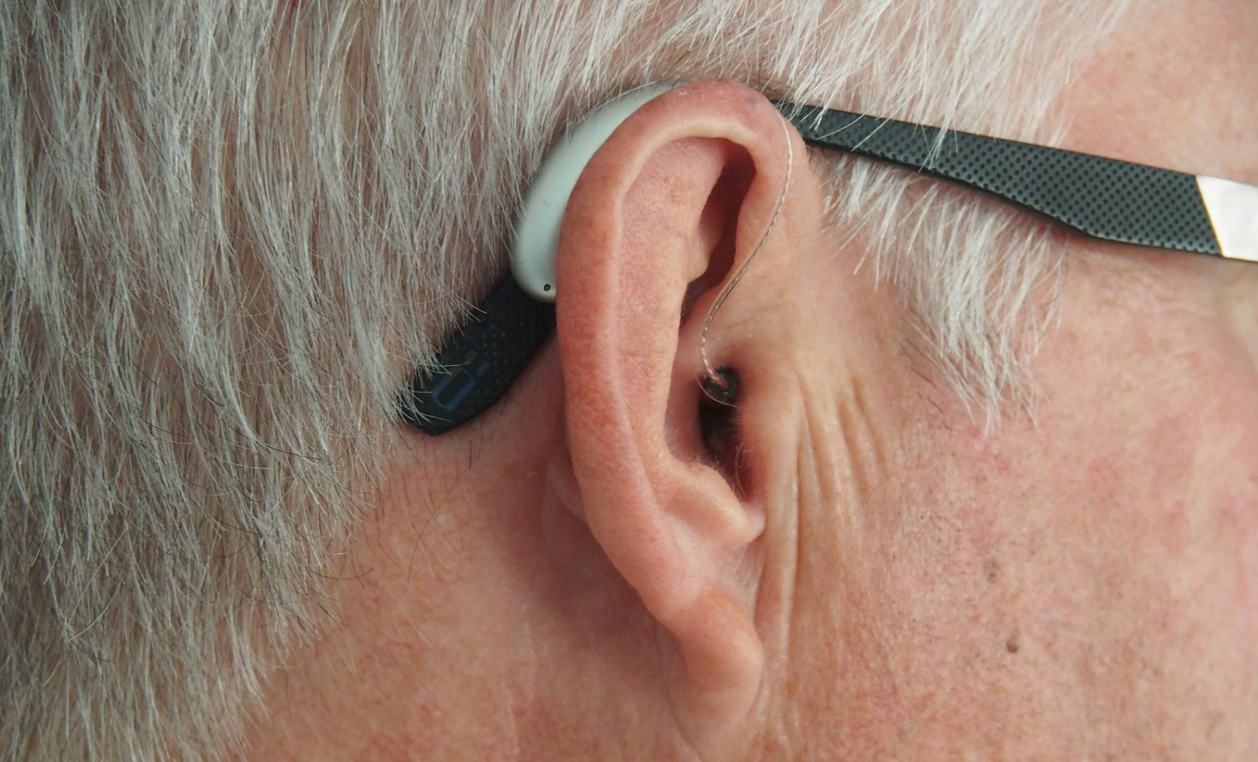 An older man wearing glasses uses a hearing aid.