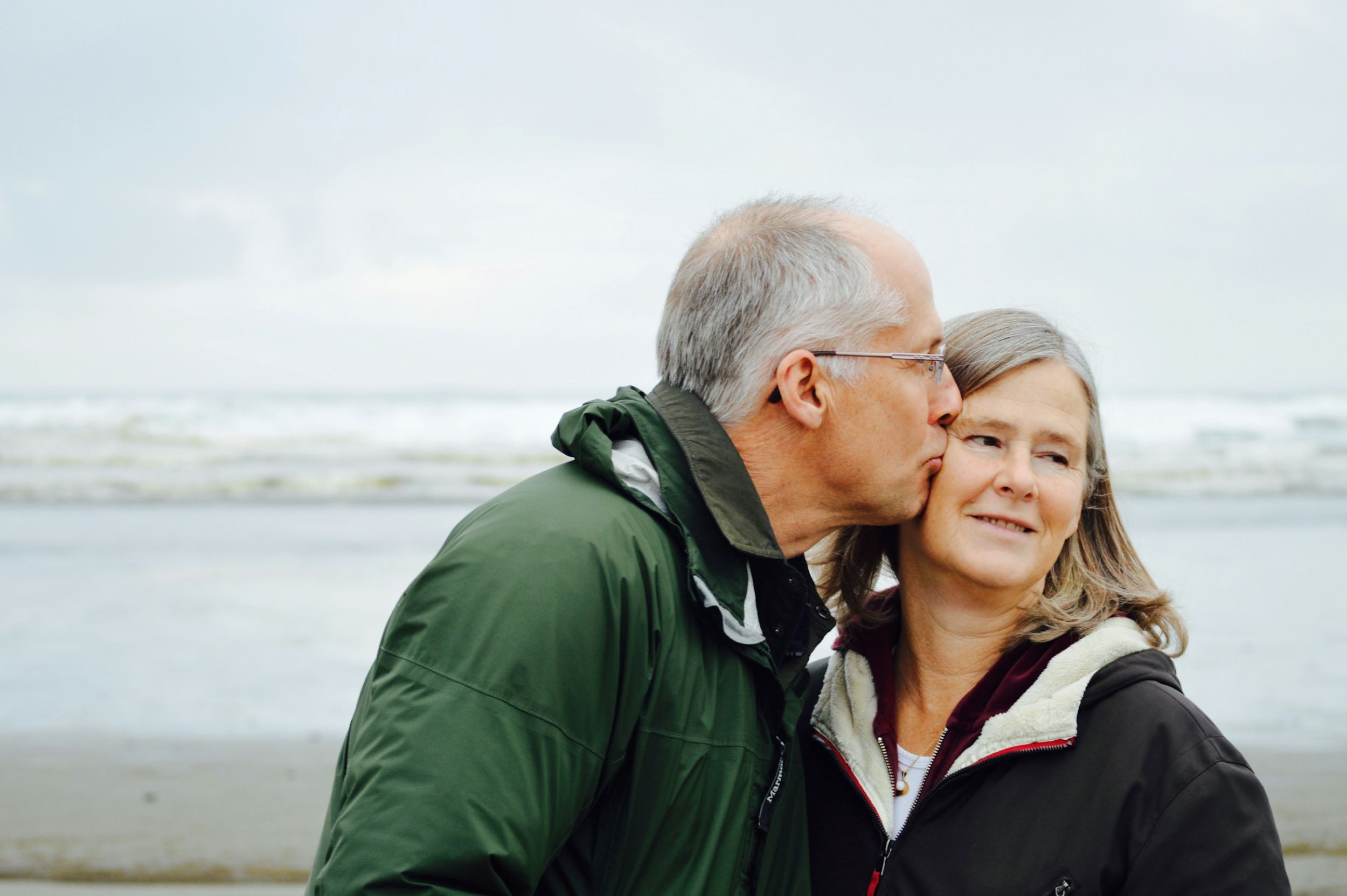 A senior man shares a tender kiss with his wife, with the ocean and sandy beach stretching out behind them.