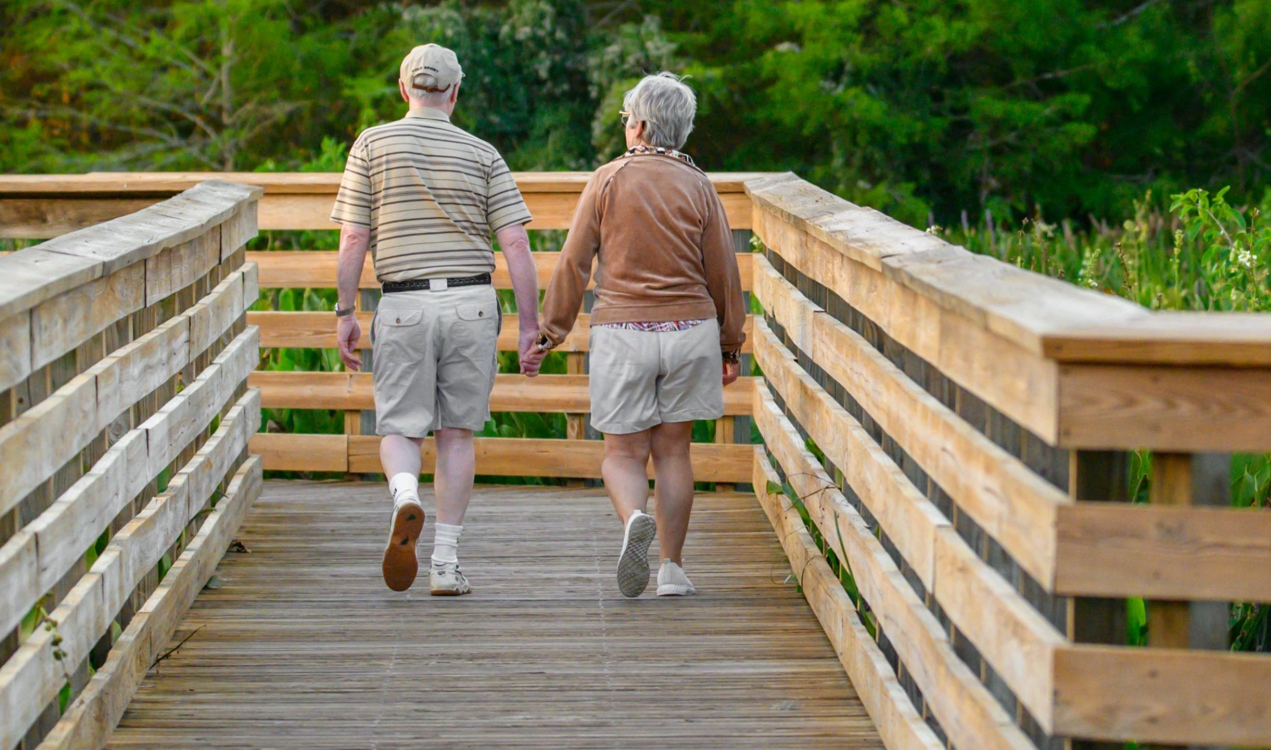 An elderly couple strolls hand in hand along a scenic boardwalk.