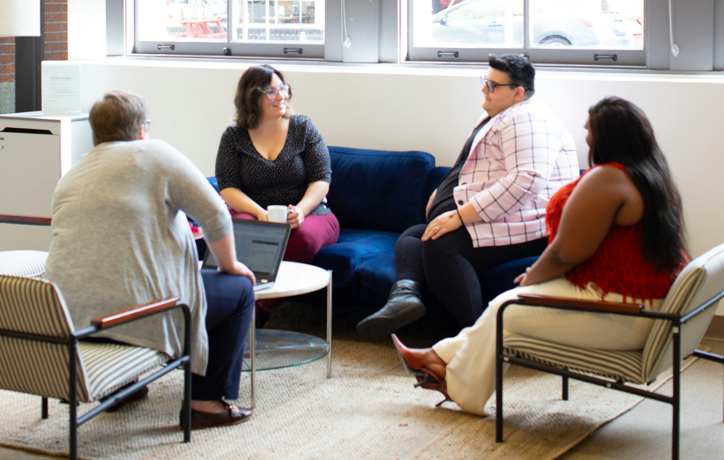 Men and women sit in a room with white chairs and speak amongst each other.