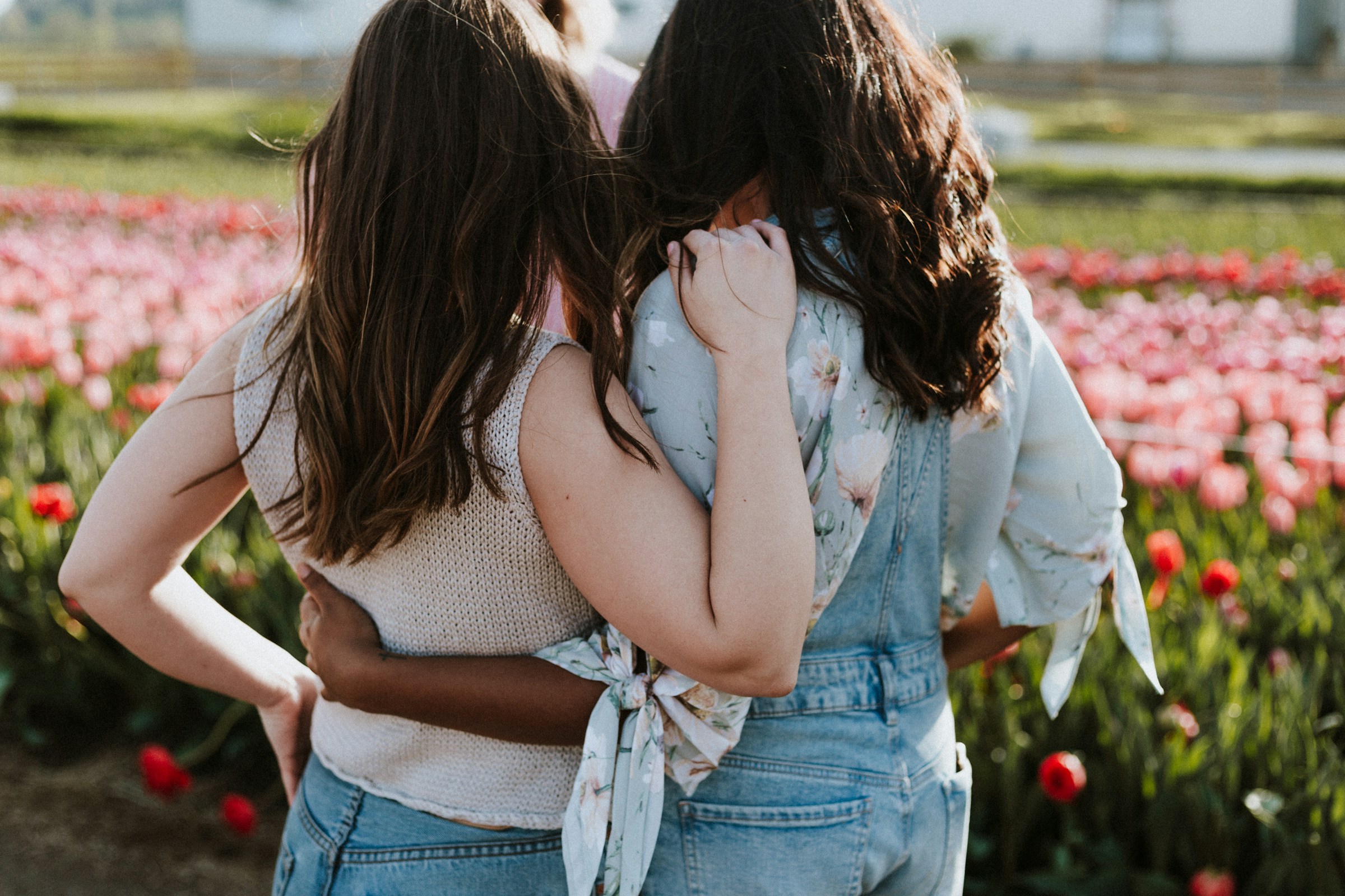 Two women share an embrace while standing in front of a valley filled with pink blossoms.