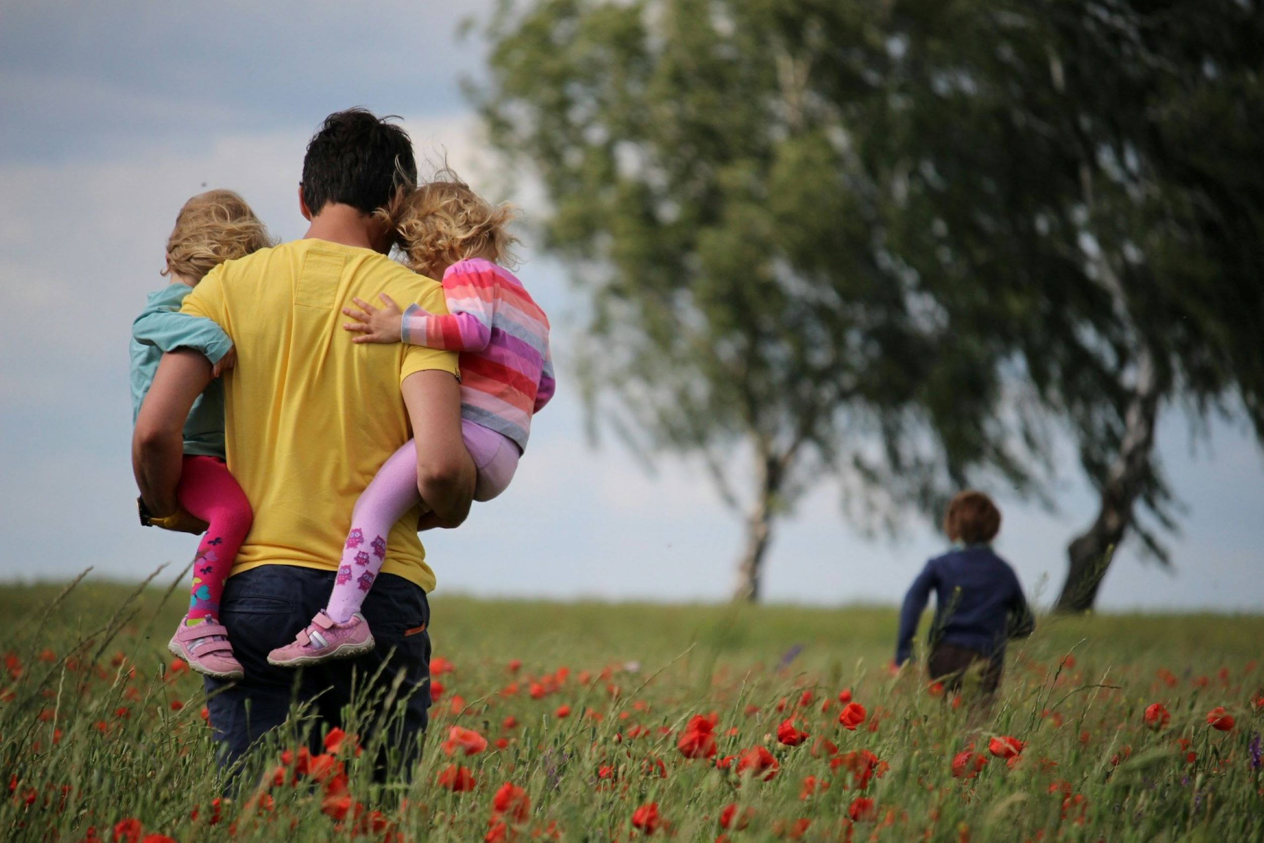 A father carries his two daughters and walks toward an oversized tree on a sunny day. 