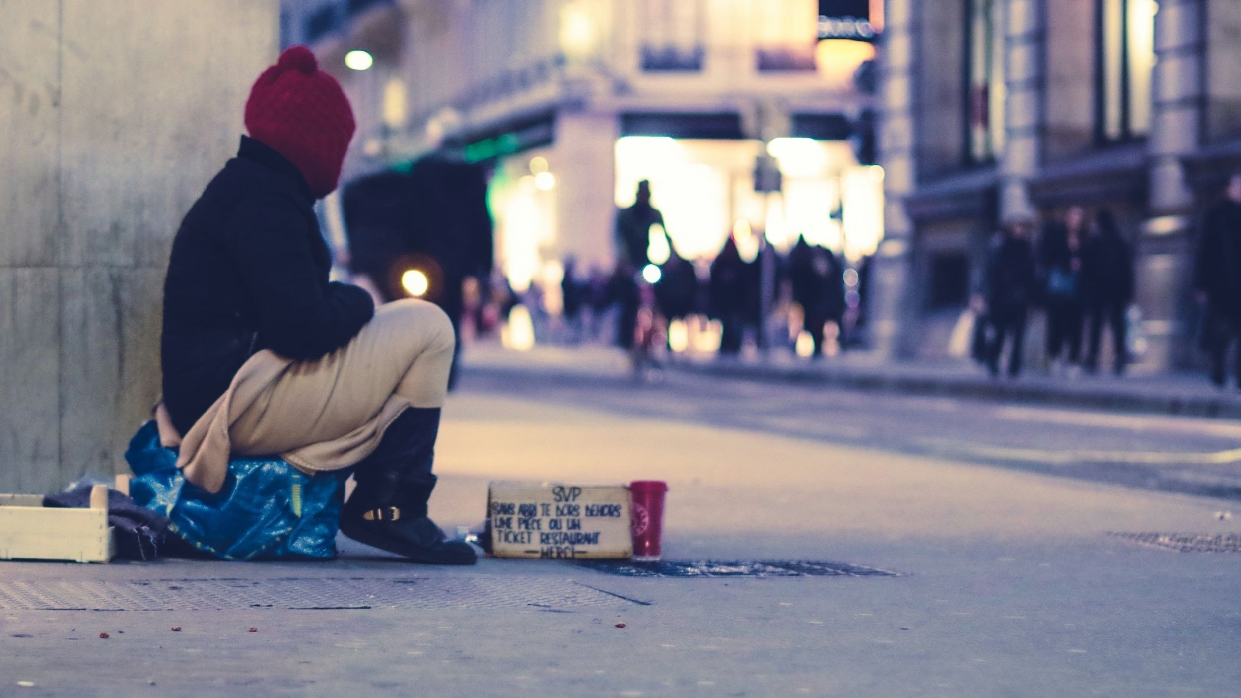 A homeless individual sits on a small crate, watching people pass by.