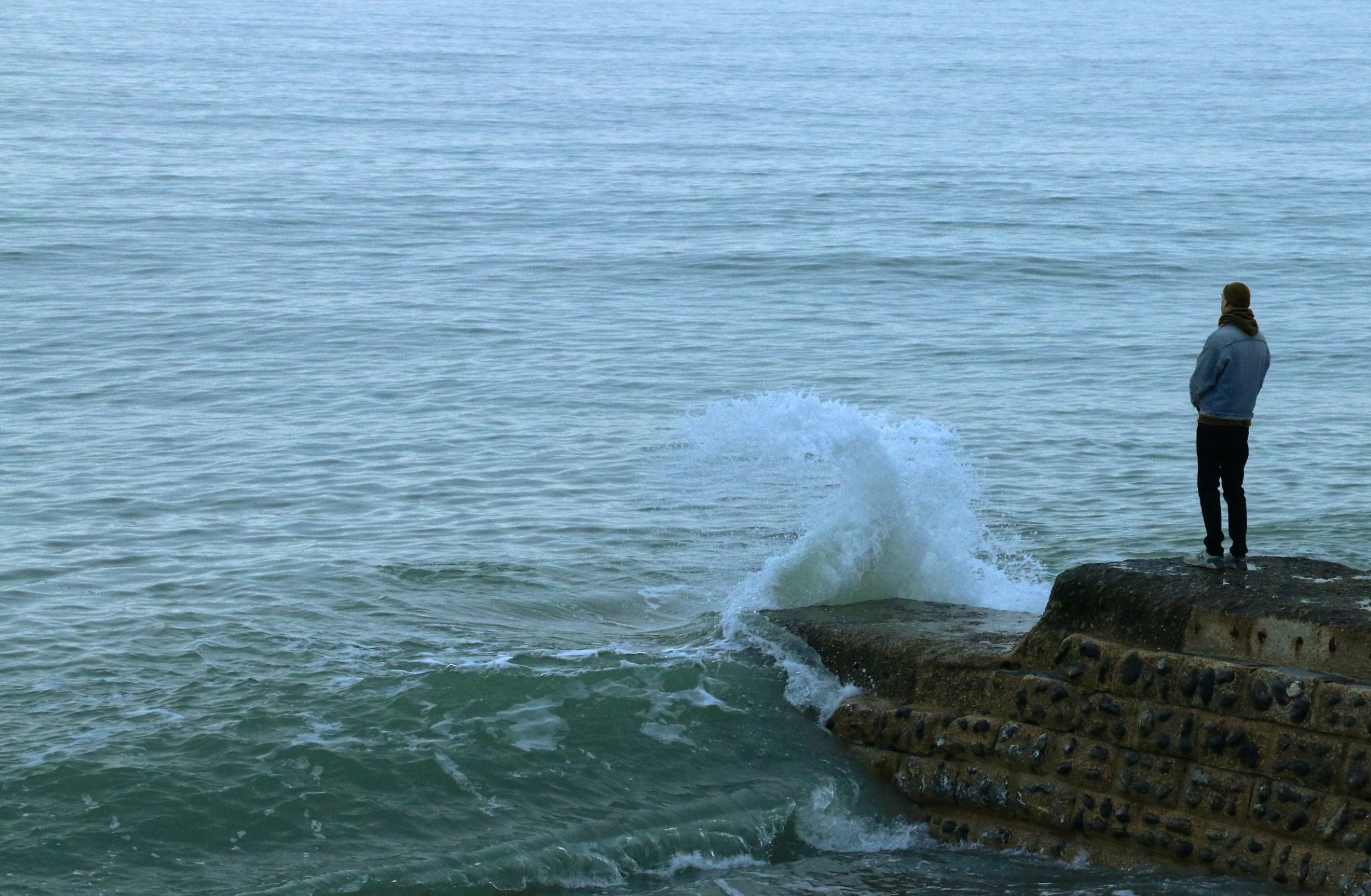 A man stands on the edge of a rock and stares at waves thrashing against the shore.